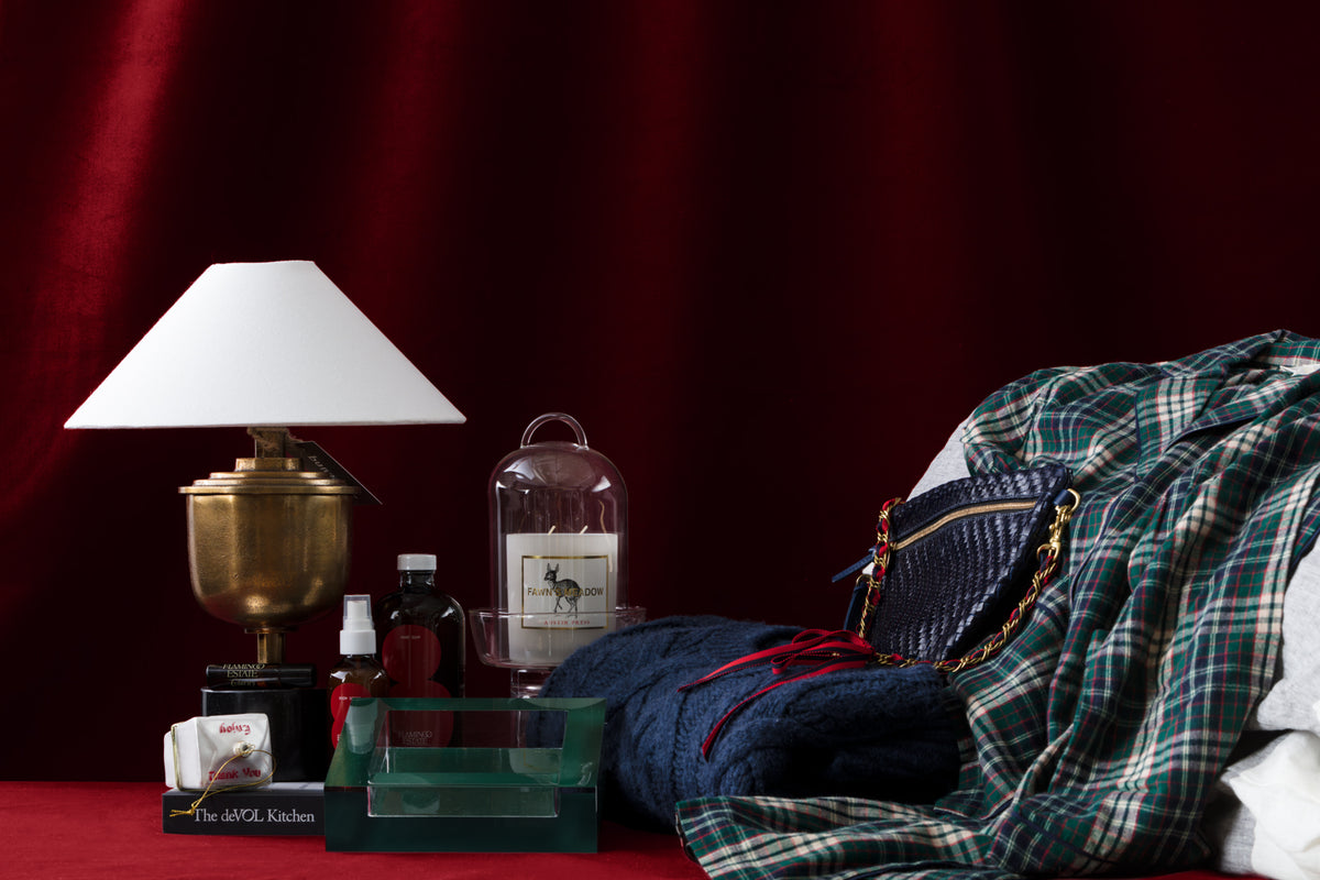 A brass lamp with a white shade, toiletries, a candle, books, a green tray, a blue textured bag with a chain strap, and plaid fabric are arranged on a dark red background.