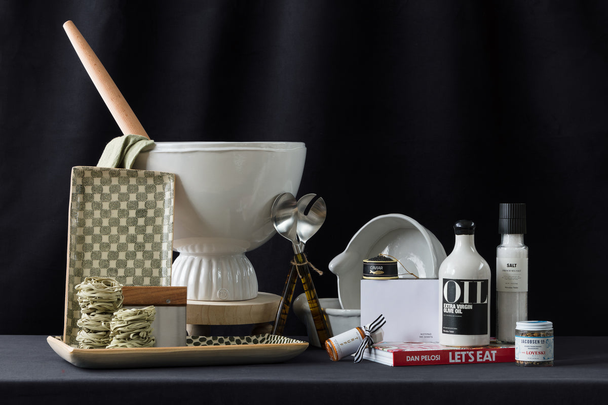 Still life featuring kitchen items: a large white mortar with pestle, oil, salt, pepper grinders, a bowl, stacked green crackers, utensils, a book, canned food, and a woven tray, set against a black background.