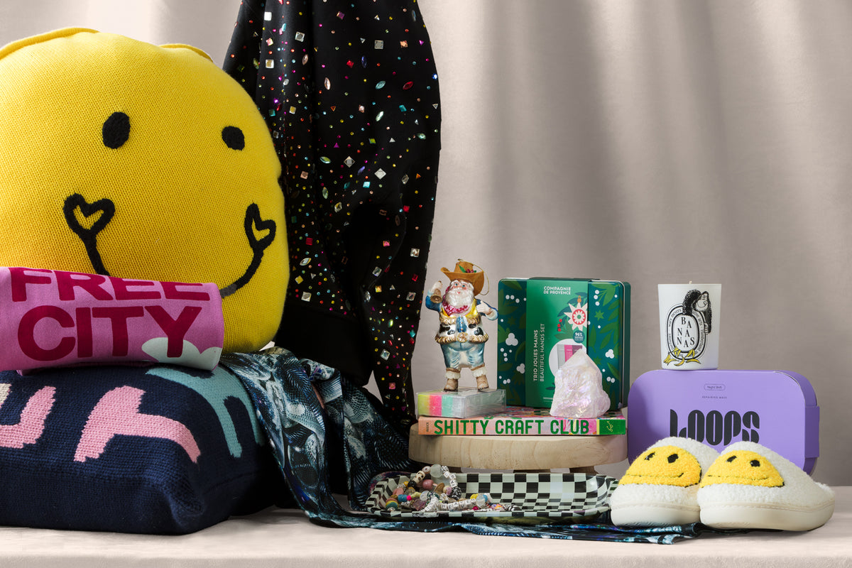 A colorful assortment of gifts includes a large yellow smiley-face pillow, cozy slippers, sweaters, a scarf, books, a figurine, a decorated tumbler, and a purple speaker, all arranged on a beige surface.