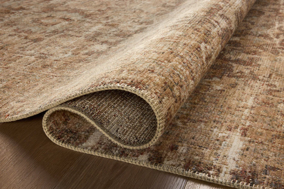 A close-up of a beige and brown patterned rug with one corner slightly curled up, revealing the texture and stitching along the edge, resting on a wooden floor.