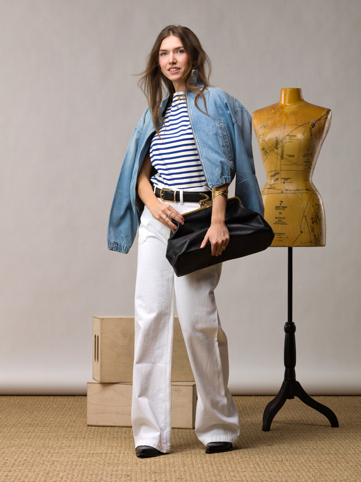 A woman wearing a striped shirt, denim jacket, and white pants holds a black clutch. She stands beside a dress form and wooden boxes against a neutral background.
