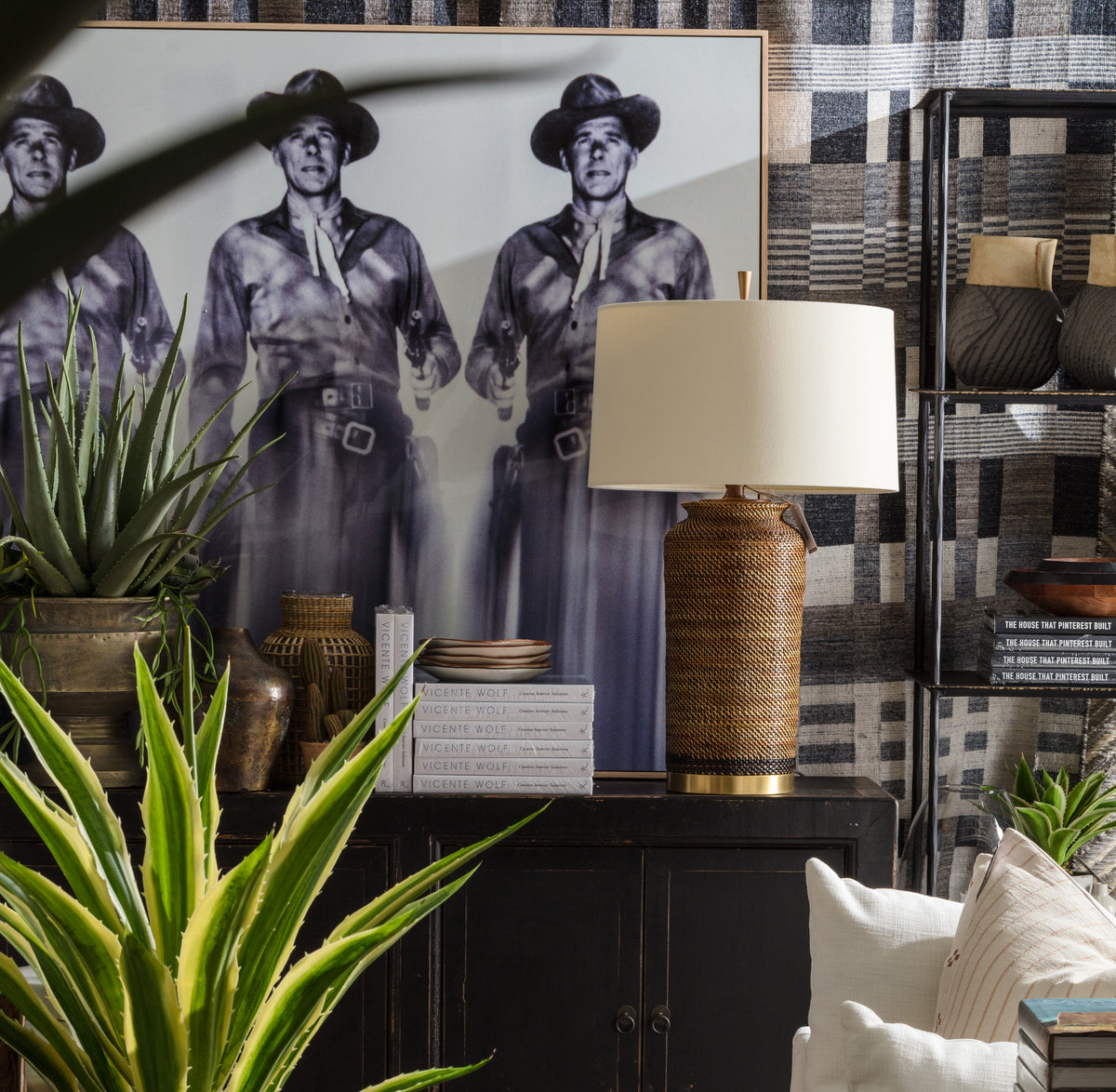 A stylish living room with potted plants, a woven table lamp, stacked books, a large black-and-white photo of three uniformed men, and patterned shelves in the background.