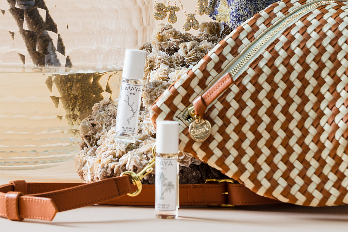 A woven bag with leather straps is placed next to two small bottles of "Maya" perfume. Behind them are dried flowers and a clear glass vase. A gold zipper and metal tag add detail to the bag.