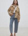 A woman stands against a plain background wearing the SUNCOO Elliott Jacket, styled over a white and brown polka dot shirt, blue high-waisted jeans, and black heeled shoes. Her hair is loose and one hand is in her pocket.