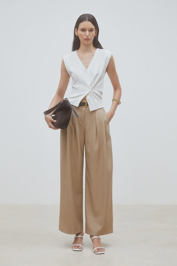 A woman poses against a plain background wearing the SUNCOO Gilem Vest layered over a sleeveless white V-neck top, high-waisted wide-leg beige trousers, white sandals, and holding a large black clutch for added elegance.
