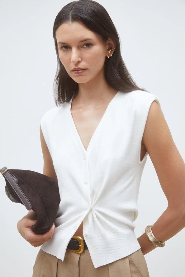A woman with long dark hair wears the SUNCOO Gilem Vest, a sleeveless white V-neck top with subtle shoulder pads, paired with beige pants, gold jewelry, and a dark brown clutch against a plain background.