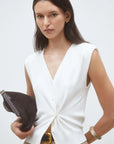 A woman with long dark hair wears the SUNCOO Gilem Vest, a sleeveless white V-neck top with subtle shoulder pads, paired with beige pants, gold jewelry, and a dark brown clutch against a plain background.