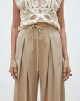A person models SUNCOO's Woven Pantalon Jeff—high-waisted, wide-leg pants with pleats and a drawstring—paired with a sleeveless, intricately crocheted cream top. Only the torso and upper legs are shown.