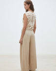 A woman stands in a minimalist, light-toned room, wearing SUNCOO's Woven Pantalon Jeff—high-waisted, wide-leg pants crafted from FSC viscose—paired with a sleeveless cream crochet top.