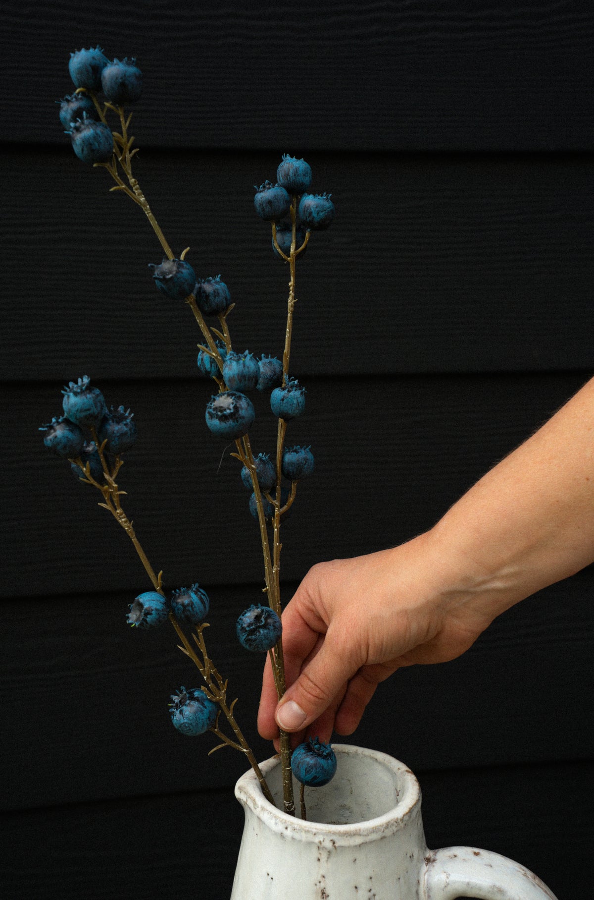A hand arranges the realistic Indaba Blueberry Branch, ideal as faux flowers or accent décor, into a white ceramic pitcher set against a dark, textured backdrop.