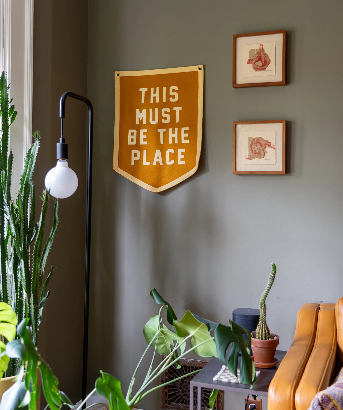 A cozy living room corner features green plants, framed wall art, and the Faire "This Must Be The Place" Camp Flag in yellow. A modern floor lamp and brown sofa add to the inviting home decor.