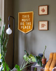 A cozy living room corner features green plants, framed wall art, and the Faire "This Must Be The Place" Camp Flag in yellow. A modern floor lamp and brown sofa add to the inviting home decor.