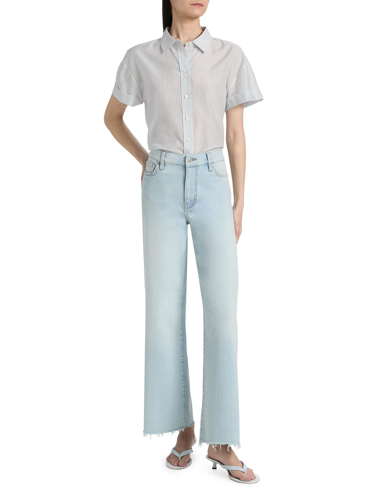 A woman wears the FRAME Boardwalk Shirt, a short-sleeved, striped cotton-silk top, tucked into high-waisted, light-wash wide-leg jeans with frayed hems and white heeled sandals, standing confidently with one hand on her hip.
