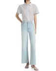 A woman wears the FRAME Boardwalk Shirt, a short-sleeved, striped cotton-silk top, tucked into high-waisted, light-wash wide-leg jeans with frayed hems and white heeled sandals, standing confidently with one hand on her hip.