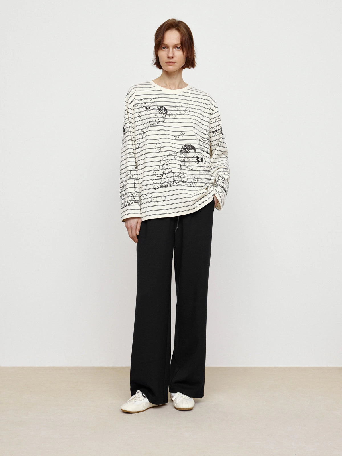 A person with short brown hair stands against a plain background, wearing the Faire Graffiti Stripe Tee paired with black wide-leg pants and white sneakers.