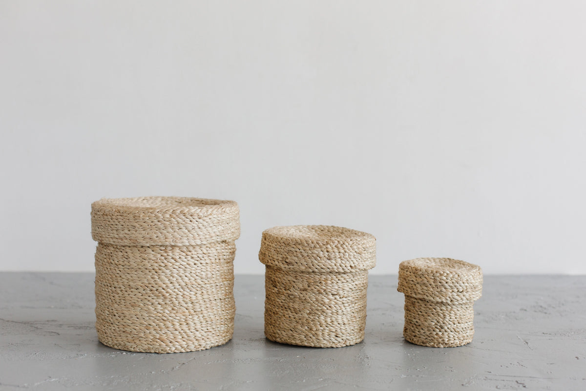 Three Faire Round Lidded Jute Baskets in assorted sizes, featuring a natural beige hue and textured finish, are lined up on a gray surface with a plain white background.