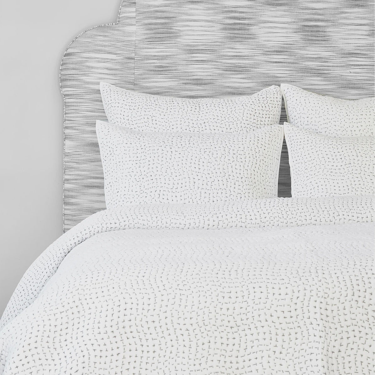 The John Robshaw Hand Stitched Coverlet King features small, subtle dashed lines reminiscent of hand-stitched quilts, atop a neatly made bed with matching pillows and a gray textured headboard for a cozy look.