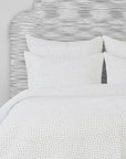 The John Robshaw Hand Stitched Coverlet King features small, subtle dashed lines reminiscent of hand-stitched quilts, atop a neatly made bed with matching pillows and a gray textured headboard for a cozy look.