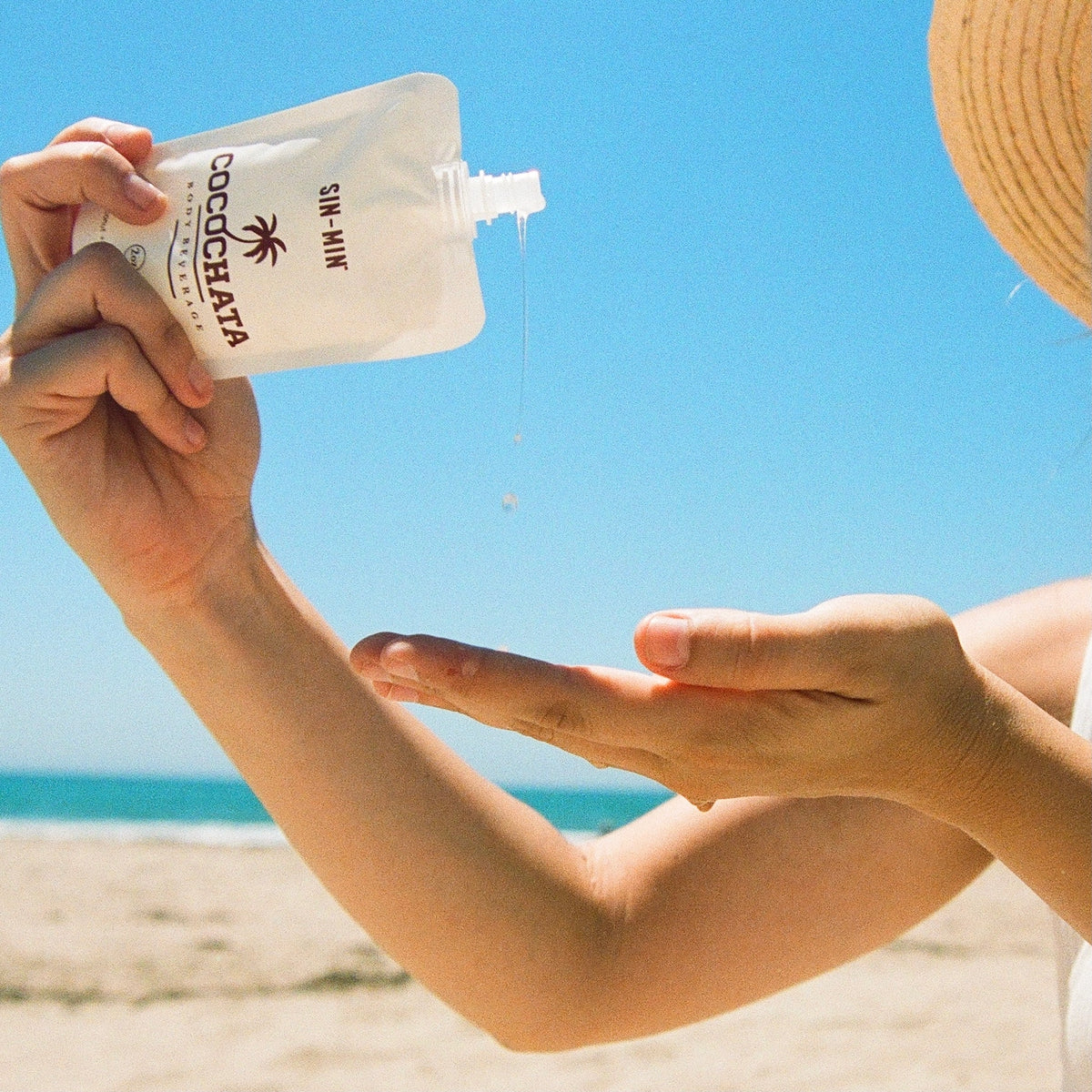 A person squeezes Faire Cocochata Body Beverage onto their hand at a sunny beach, with blue sky and sand in the background—ideal for those seeking a sensitive skin moisturizer on warm, sunny days.