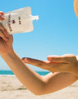 A person squeezes Faire Cocochata Body Beverage onto their hand at a sunny beach, with blue sky and sand in the background—ideal for those seeking a sensitive skin moisturizer on warm, sunny days.