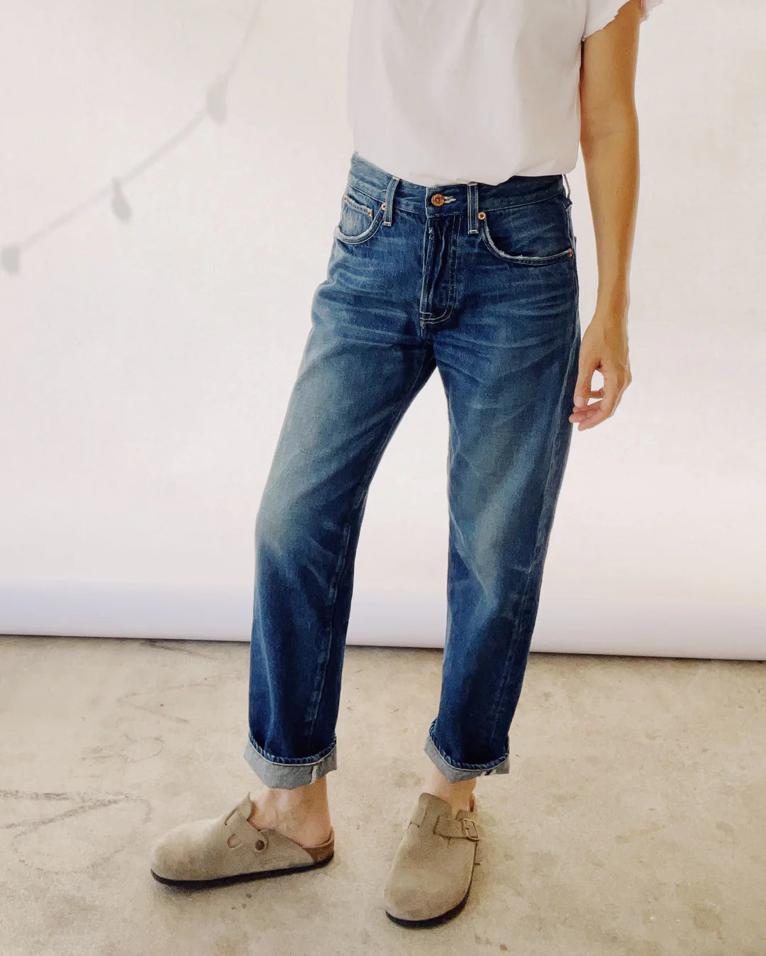 Someone stands casually wearing The Loose Crop jeans by Hey Gang, made from 100% cotton denim, a white T-shirt, and tan slip-on shoes. Captured from the shoulders down against a neutral background with faint string lights visible.