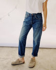 Someone stands casually wearing The Loose Crop jeans by Hey Gang, made from 100% cotton denim, a white T-shirt, and tan slip-on shoes. Captured from the shoulders down against a neutral background with faint string lights visible.