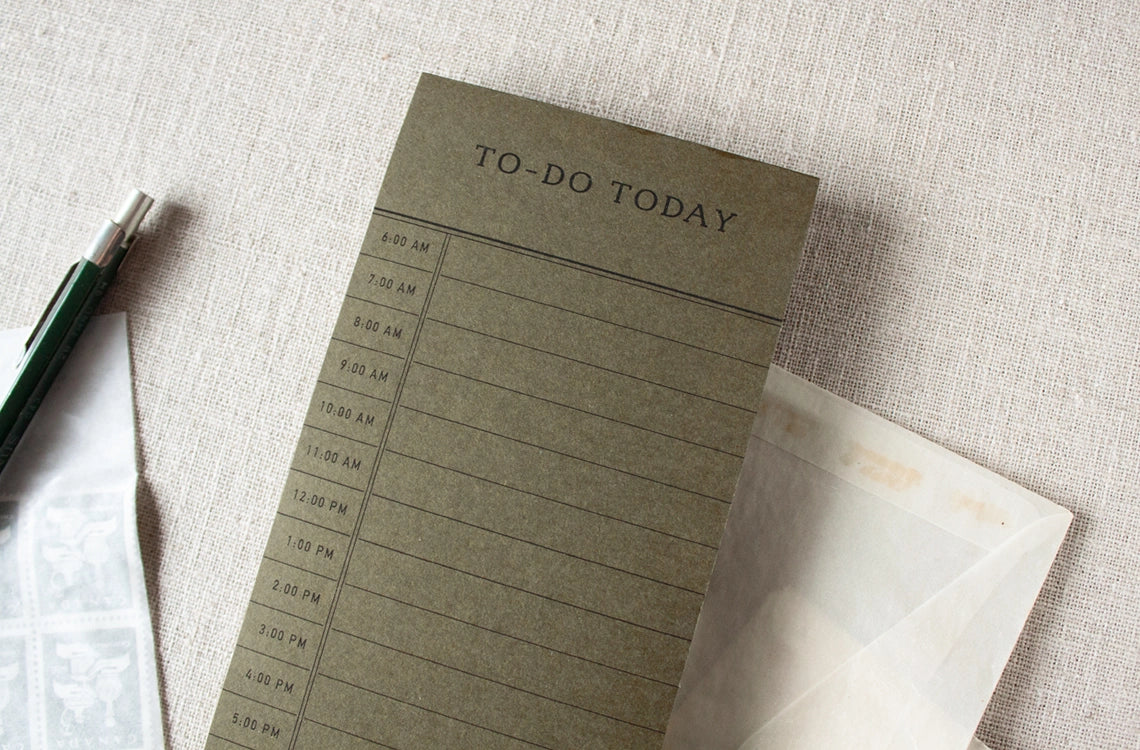The Faire To Do Today Planner is a green 50-sheet notepad with hourly slots from 9 AM to 5 PM. It sits on a light textured surface alongside a pen, postage stamps, and an open envelope.