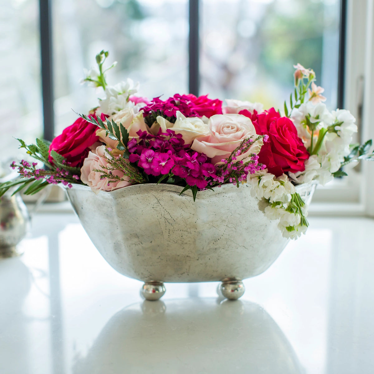 The Faire Vintage Scalloped Bowl, handmade in Italy, holds a vibrant mix of pink, red, and white roses along with assorted blooms. It rests on a white surface near a window with a softly blurred outdoor backdrop.