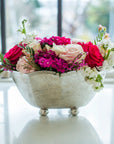 The Faire Vintage Scalloped Bowl, handmade in Italy, holds a vibrant mix of pink, red, and white roses along with assorted blooms. It rests on a white surface near a window with a softly blurred outdoor backdrop.