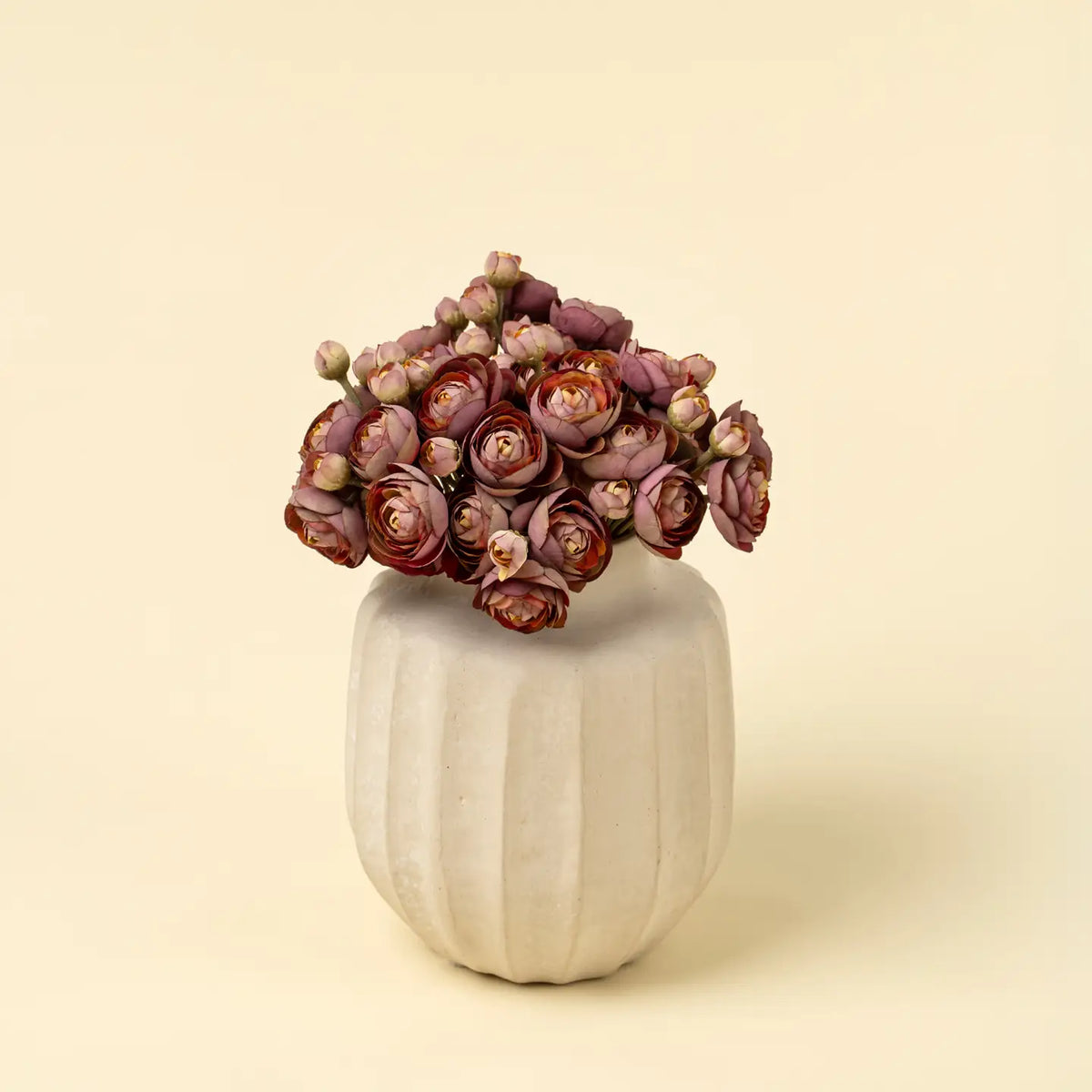 A Ranunculus Bouquet by Faire features small, pinkish-maroon flowers arranged in a white, textured ceramic vase from our Decorative Vases collection, set against a plain light beige background.