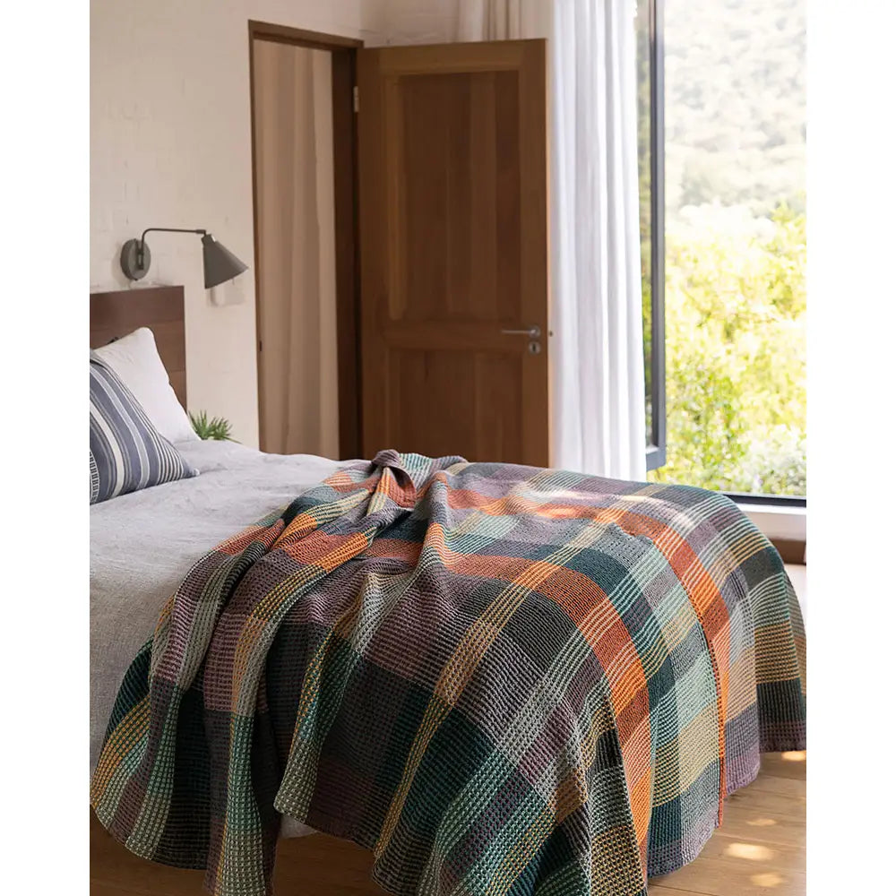 A cozy bedroom features a Faire Vrou-Vrou blanket with bold design on the bed, white linens, and a striped pillow. Sunlight pours in through an open door and large window, revealing lush greenery outside.