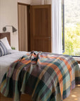 A cozy bedroom features a Faire Vrou-Vrou blanket with bold design on the bed, white linens, and a striped pillow. Sunlight pours in through an open door and large window, revealing lush greenery outside.