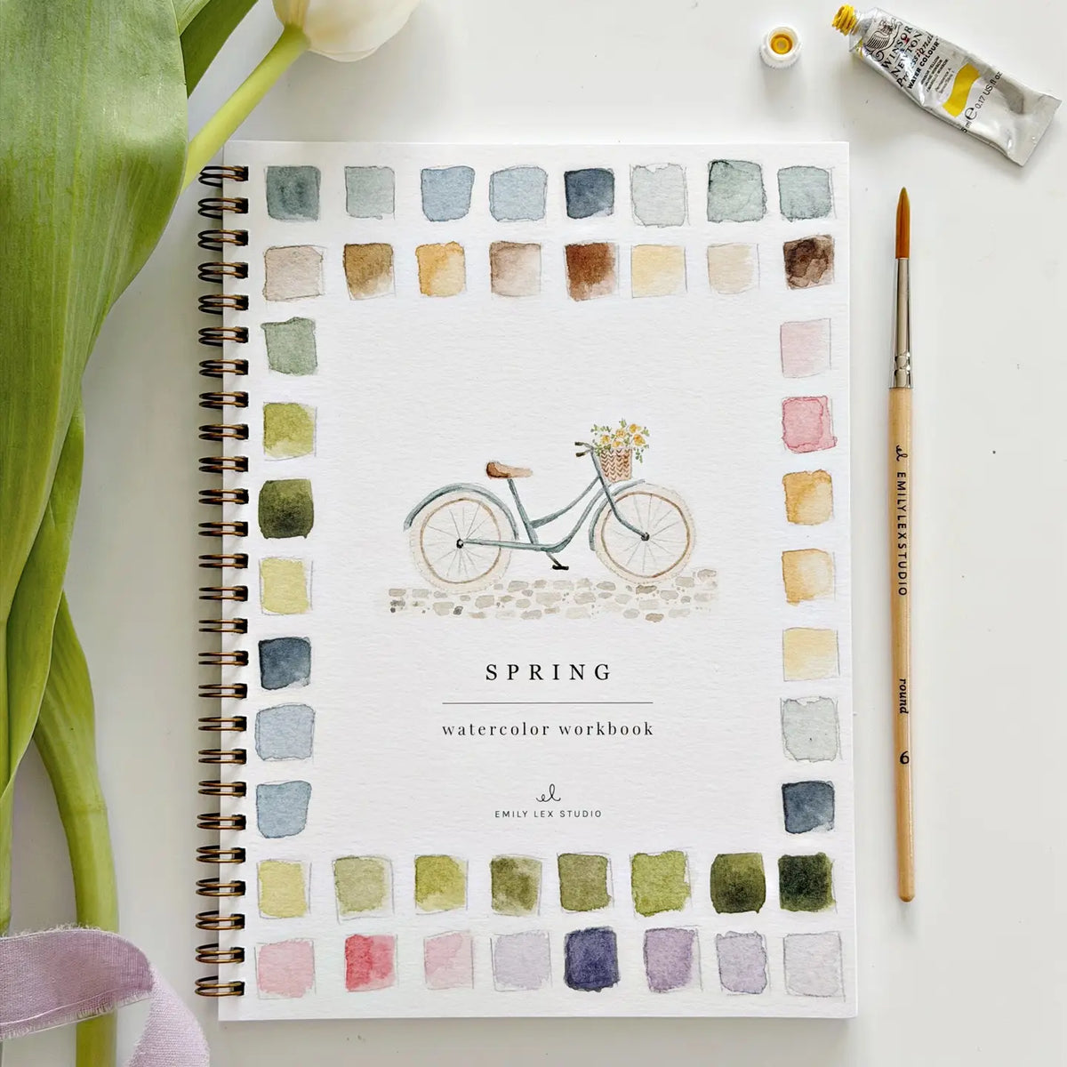 The Faire Watercolor Workbook is a spiral-bound beginner’s guide featuring a bicycle illustration and vibrant paint swatches on the cover, pictured with a paintbrush, paint tube, and green florist stems on a white surface.