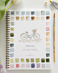The Faire Watercolor Workbook is a spiral-bound beginner’s guide featuring a bicycle illustration and vibrant paint swatches on the cover, pictured with a paintbrush, paint tube, and green florist stems on a white surface.