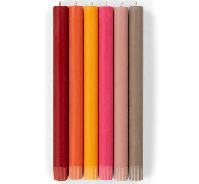 Six tall, cylindrical Faire Warm Colours Dinner Candles S/2—dark red, red, orange, yellow, pink, and taupe with unlit wicks—are hand poured and Fair Trade. The candles are shown in a row against a white background.
