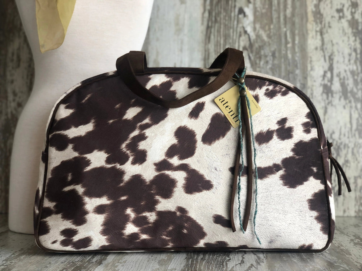 The Faire Oakley Overnighter- Faux Cowhide is a vegan travel bag featuring a brown and white cowhide pattern, dark handles, and an "alema" tag with ribbon, displayed before a light mannequin and rustic wooden background.