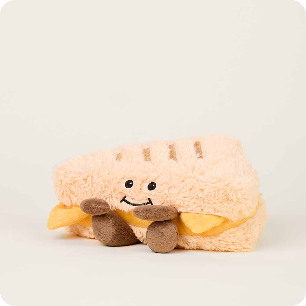 The Faire Grilled Cheese Warmies is a microwavable plush shaped like a smiling sandwich with brown limbs and yellow felt cheese, perfect for cozy comfort on any light background.
