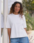 Wavy-haired woman in a Frank & Eileen Perry Crew SS Tee and denim shorts smiles casually by a white wall.