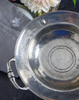 The Faire Vintage 1795 Pedestal Bowl with handles and stamped markings rests on a dark surface beside a folded embroidered gray napkin and a vase holding a white dahlia flower.