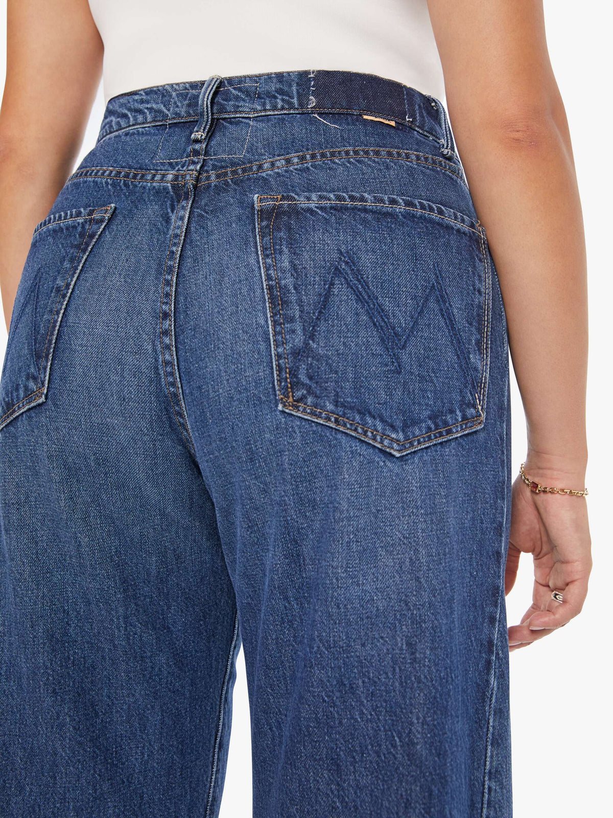 A person wears Mother’s The Half-Pipe Ankle jeans—super high-waisted, wide-leg denim in dark blue with a stitched "M" on the back pocket—and a white top; their right arm shows a delicate bracelet.
