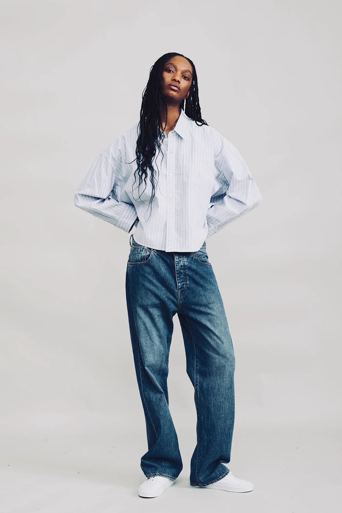 Blue striped cropped oxford shirt with loose blue jeans and white sneakers, styled by Denimist on a plain background.