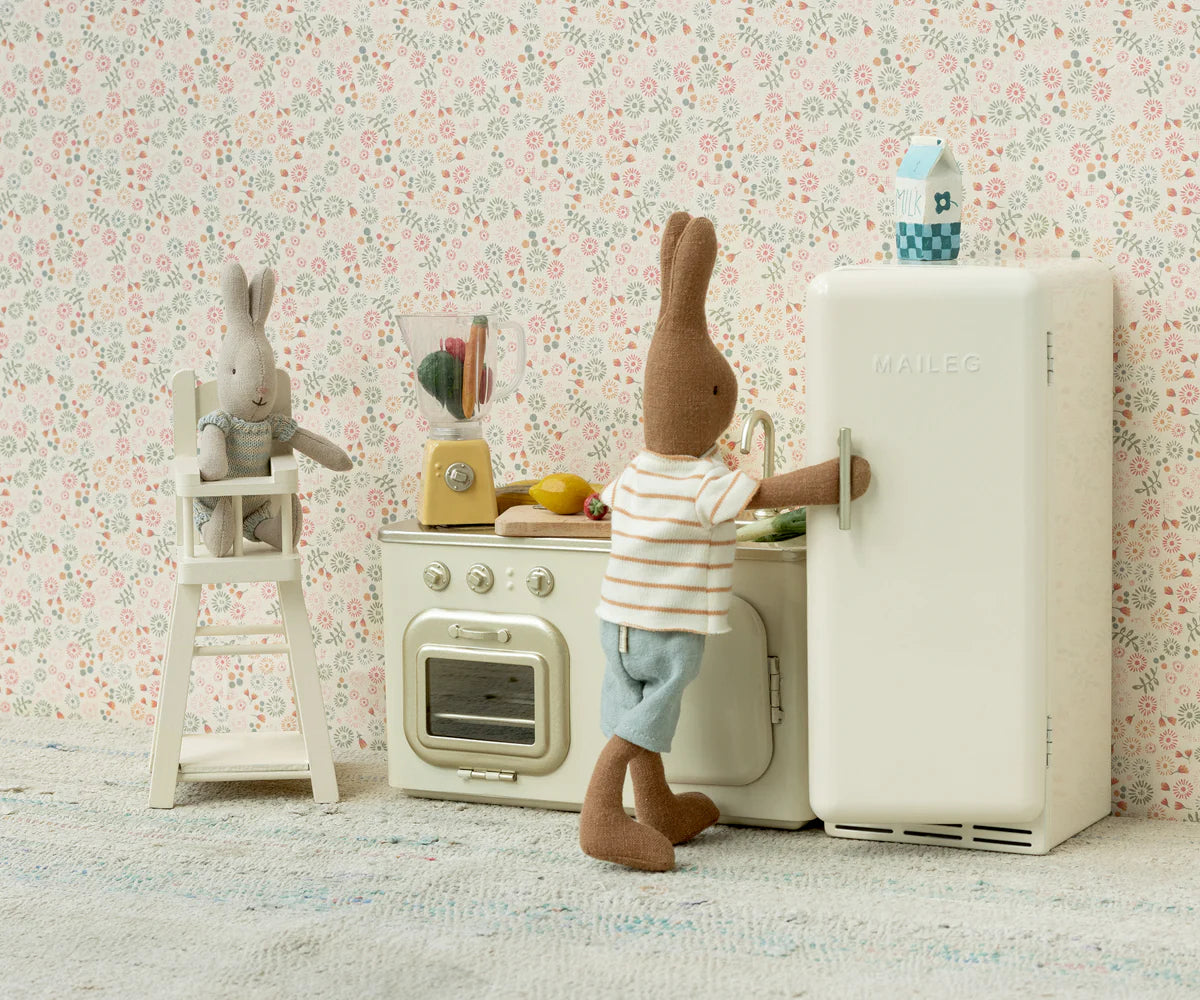 Two Maileg bunny toys play in a miniature kitchen: one is by the fridge, the other sits in a high chair near a stove with the Maileg Blender and tiny food, all set against charming floral wallpaper.
