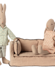 Two fabric bunny dolls sit near a Maileg Couch, Miniature by Maileg: one in pale green pajamas stands beside it, while the other in a pink dress sits on the sofa. Both are set against a plain white background.