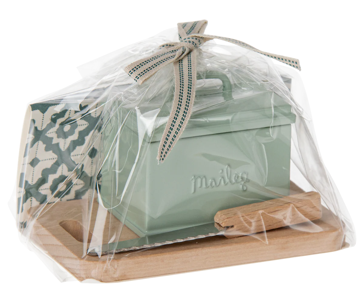 A Maileg Bread box w. utensils by Maileg is wrapped in clear plastic with a ribbon, displayed on a wooden tray next to a patterned card and a small green tin mailbox—charming accessories for any dollhouse.