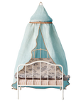 The Maileg Canopy Bed by Maileg features a small white metal frame with patterned bedding and a light blue fabric canopy trimmed in gold, draped from an overhead ring—a charming touch for any bedroom decor.