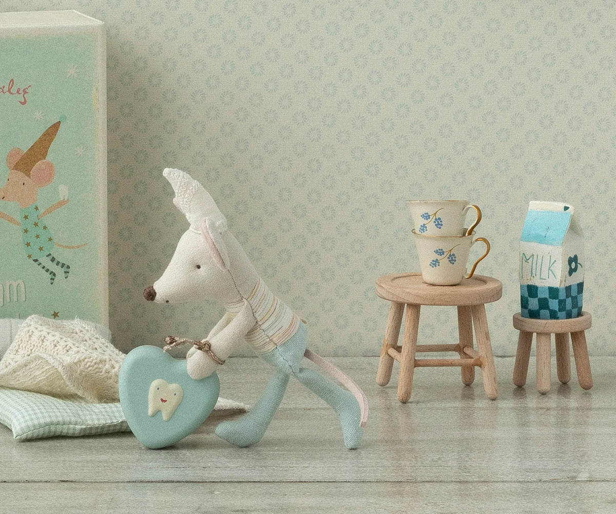 A small fabric mouse in blue pants holds a blue heart-shaped box with a tooth image. Next to it are two stacked teacups on the Maileg Table & Stool by Maileg, with a milk carton and patterned wallpaper in the background.