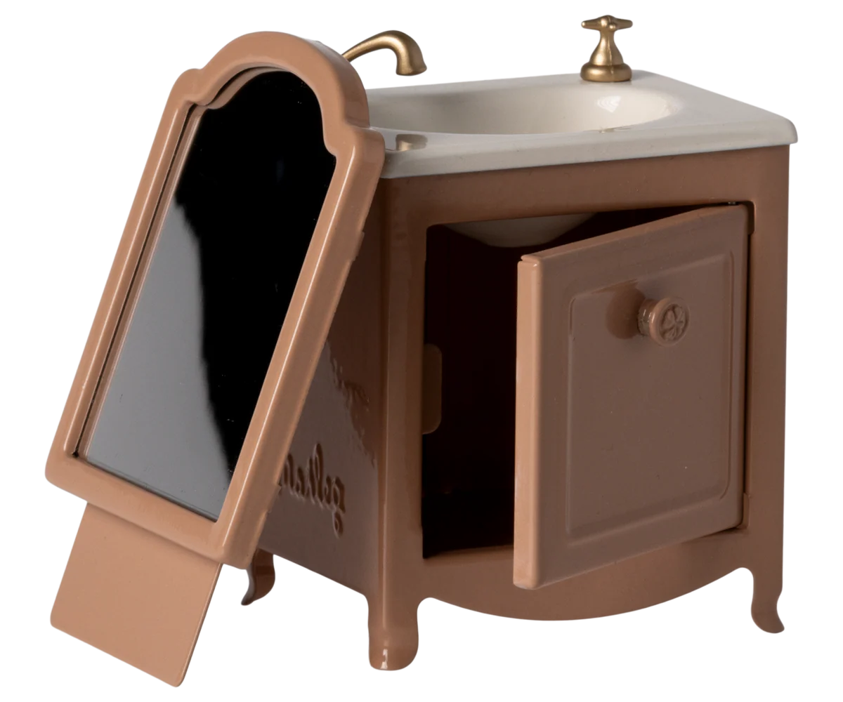 The Maileg Sink Dresser is a small brown toy vanity featuring a white bathroom sink, gold faucet, side mirror on the left, ramp at the base, and an open cabinet door—ideal as dollhouse furniture.
