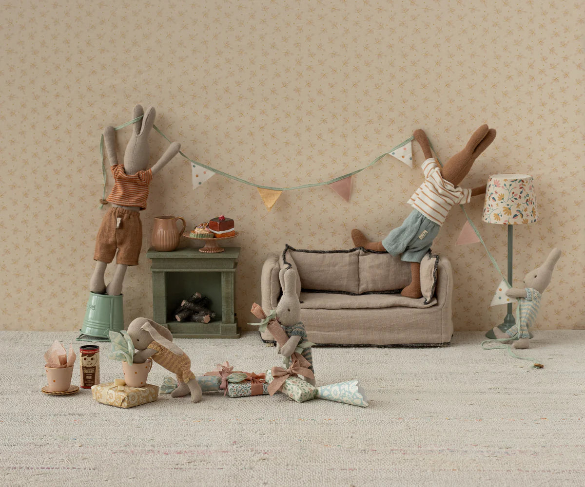 Four Maileg stuffed bunnies decorate a cozy dollhouse room for a party. Two hang bunting above the couch, while two more play and unwrap gifts and the Maileg Cakes Display set on the floor.