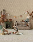 Four Maileg stuffed bunnies decorate a cozy dollhouse room for a party. Two hang bunting above the couch, while two more play and unwrap gifts and the Maileg Cakes Display set on the floor.