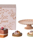 The Maileg Cakes Display by Maileg features a decorative “Petit fours” box, a pink scalloped cake stand, and three dollhouse pastries artfully arranged on a white background.